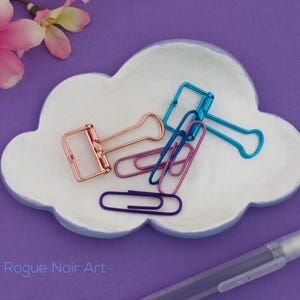May include: A white cloud-shaped dish with a blue rim holds various office supplies. Copper, blue, and purple binder clips and paper clips are arranged on the dish. A clear pen rests on the purple surface. The text "Rogue Noir Art" is visible.