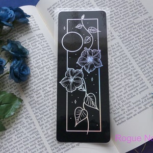 May include: A black and white bookmark with a floral design and a moon. The bookmark has a holographic finish and the text "Rogue Noir Art" is printed at the bottom.