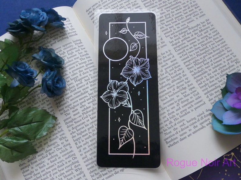 May include: A black and white bookmark with a floral design and a moon. The bookmark has a holographic finish and the text "Rogue Noir Art" is printed at the bottom.
