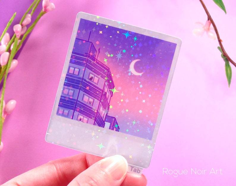 May include: A holographic sticker featuring a purple and pink cityscape with a crescent moon and stars. The sticker has a white border and the text "Tab" and "Rogue Noir Art" are visible.