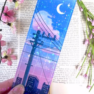 May include: A holographic bookmark featuring a cityscape at night with a crescent moon and stars in the sky. The cityscape is outlined in pink and blue, with a power line running through the center of the image.