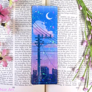 May include: A holographic bookmark with a cityscape silhouette, a crescent moon, and stars against a blue and pink sky. The bookmark has a glittery, iridescent finish.