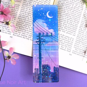 May include: A holographic bookmark with a cityscape silhouette, a crescent moon, and stars against a pink and blue gradient sky. The bookmark has a grid pattern and glitter accents.