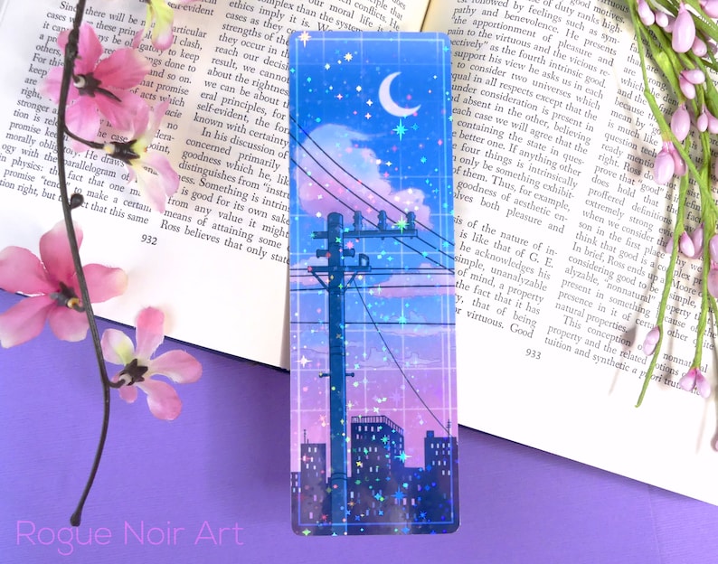 May include: A holographic bookmark with a cityscape silhouette, a crescent moon, and stars against a pink and blue gradient sky. The bookmark has a grid pattern and glitter accents.