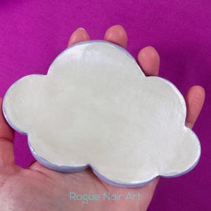 May include: A cloud-shaped ceramic dish with a pale blue rim and a white interior. The dish is held in a hand against a purple background. The text "Rogue Noir Art" is visible in the bottom right corner.