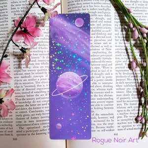May include: A purple and pink holographic bookmark with a galaxy design featuring three planets and stars. The bookmark has a holographic finish that shimmers in different colors.
