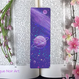 May include: A purple and blue bookmark with a galaxy design. The bookmark features a large planet with a ring, a smaller planet, and a scattering of stars. The bookmark is perfect for adding a touch of whimsy to any book.