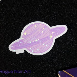 K&ouml;nnte beinhalten: Ein lila und wei&szlig;er Aufkleber mit einem Planeten mit Ringen und Sternen. Der Aufkleber hat eine holografische Oberfl&auml;che.