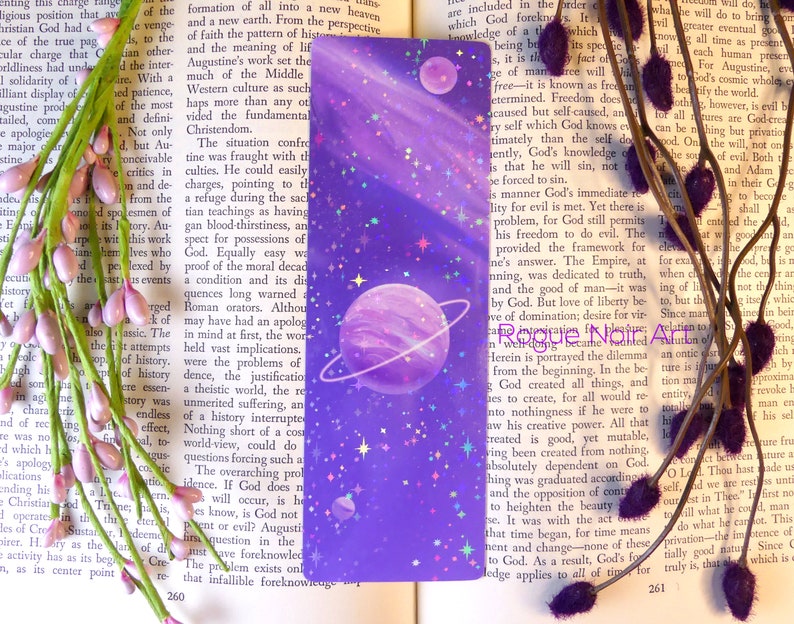 May include: A purple and pink bookmark with a galaxy design. The bookmark features a planet with rings, stars, and a holographic finish.