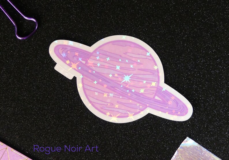 K&ouml;nnte beinhalten: Ein holografischer Aufkleber mit einem rosa Planeten mit einem Ring darum. Der Planet ist mit kleinen wei&szlig;en Sternen bedeckt. Der Aufkleber befindet sich auf einem schwarzen Hintergrund.