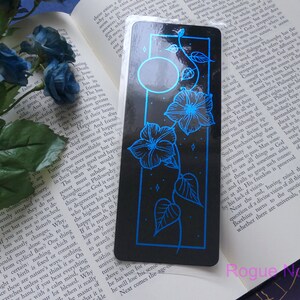 May include: A black bookmark with a blue floral design and a moon. The flowers are stylized morning glories with a vine and leaves. The bookmark has a glossy finish.