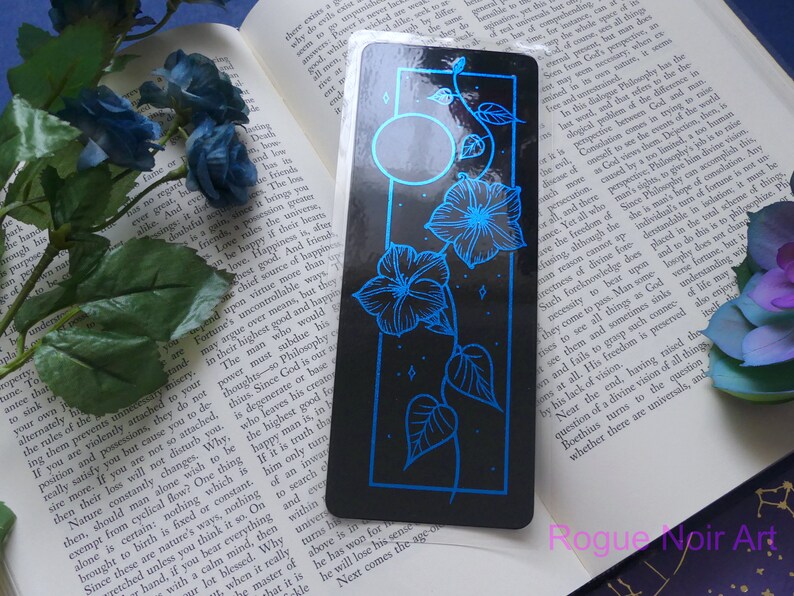 May include: A black bookmark with a blue floral design and a moon. The flowers are stylized morning glories with a vine and leaves. The bookmark has a glossy finish.