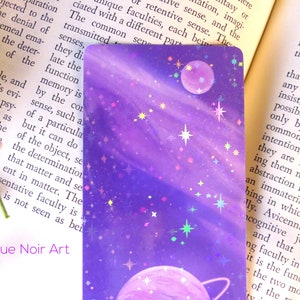May include: A purple and pink galaxy-themed bookmark with a holographic finish. The bookmark features two planets and stars. The text "Rogue Noir Art" is printed at the bottom of the bookmark.