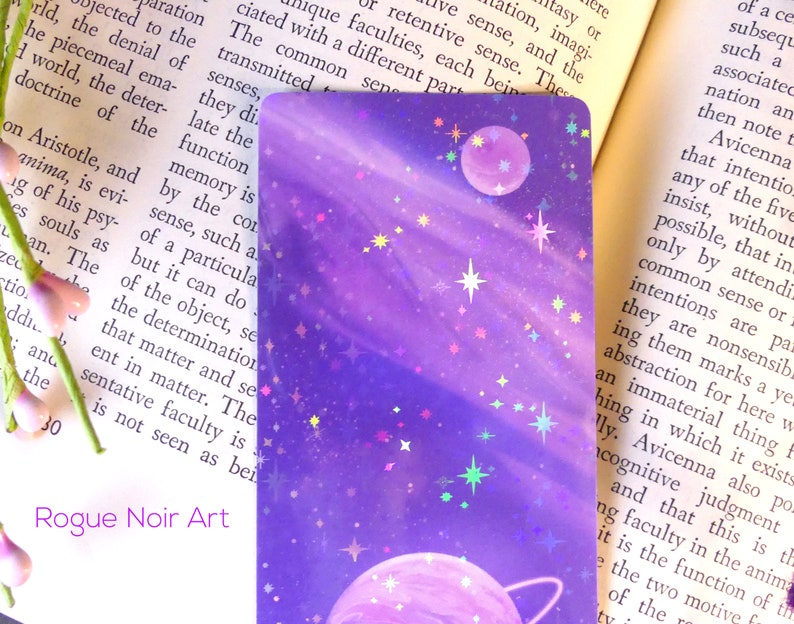 May include: A purple and pink galaxy-themed bookmark with a holographic finish. The bookmark features two planets and stars. The text "Rogue Noir Art" is printed at the bottom of the bookmark.