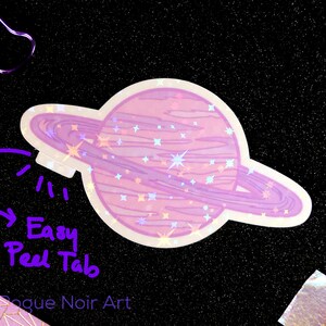K&ouml;nnte beinhalten: Ein holografischer Aufkleber mit einem rosafarbenen Planeten mit einem Ring darum. Der Planet hat eine glitzernde Textur und ist von Sternen umgeben. Der Aufkleber hat eine leicht abziehbare Lasche.
