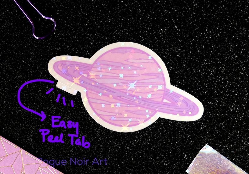 K&ouml;nnte beinhalten: Ein holografischer Aufkleber mit einem rosafarbenen Planeten mit einem Ring darum. Der Planet hat eine glitzernde Textur und ist von Sternen umgeben. Der Aufkleber hat eine leicht abziehbare Lasche.
