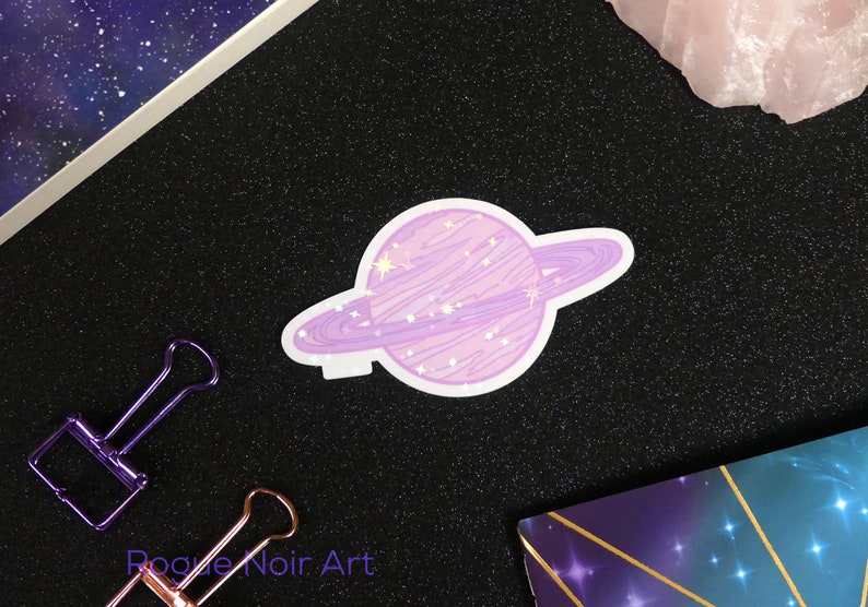 K&ouml;nnte beinhalten: Ein lila-wei&szlig;er Planetenaufkleber mit einem Ring darum. Der Planet hat ein wirbelndes Muster und wei&szlig;e Sterne darauf. Der Aufkleber befindet sich auf einem schwarzen Hintergrund.