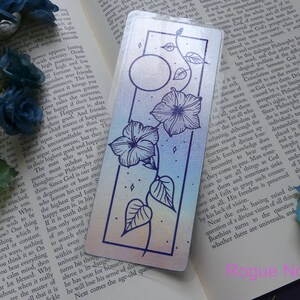 May include: A holographic bookmark with a rainbow gradient background featuring a black line drawing of a vine with two flowers and a moon. The bookmark has a silver border.