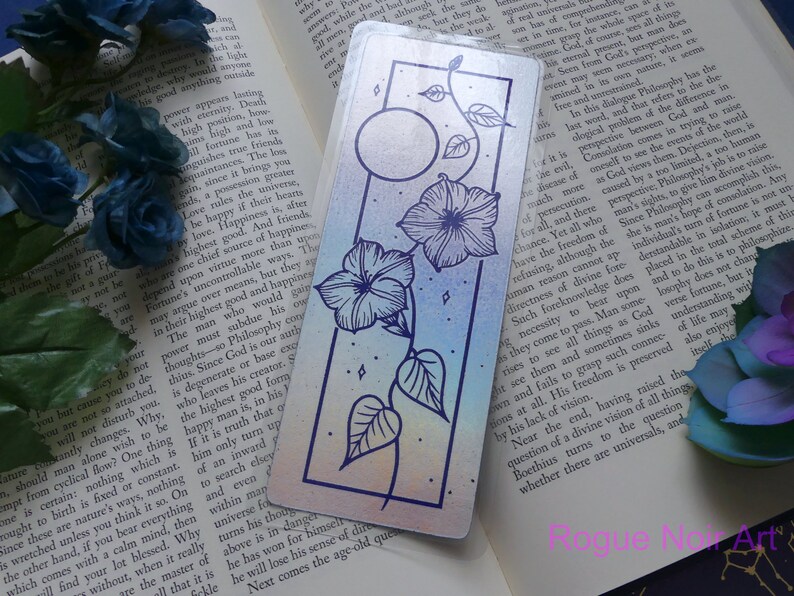 May include: A holographic bookmark with a rainbow gradient background featuring a black line drawing of a vine with two flowers and a moon. The bookmark has a silver border.