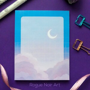 May include: A blue and white notepad with a crescent moon and clouds design. The notepad has a grid pattern and the text "Rogue Noir Art" at the bottom.