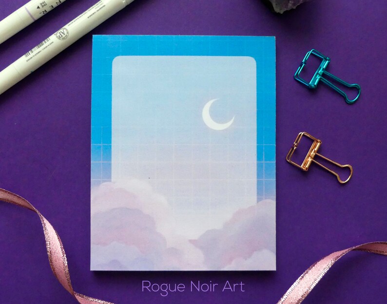May include: A blue and white notepad with a crescent moon and clouds design. The notepad has a grid pattern and the text "Rogue Noir Art" at the bottom.