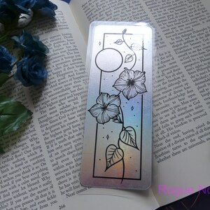 May include: A holographic bookmark with a black and white illustration of a moon, flowers, and vines. The background is a rainbow gradient.