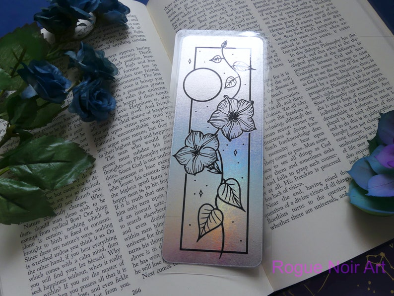 May include: A holographic bookmark with a black and white illustration of a moon, flowers, and vines. The background is a rainbow gradient.