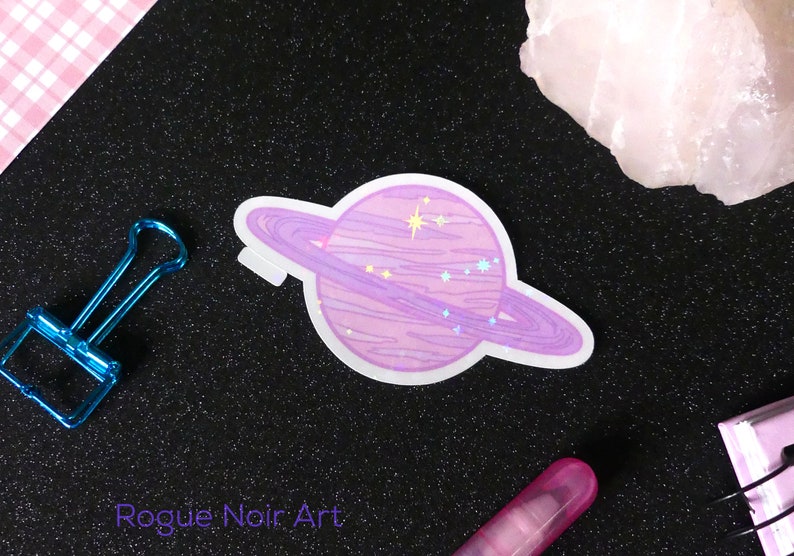 K&ouml;nnte beinhalten: Ein rosa und lilafarbener Planetenaufkleber mit einem Ring darum. Der Planet hat eine glitzernde Oberfl&auml;che und ist von Sternen umgeben. Der Aufkleber befindet sich auf einem schwarzen Hintergrund.
