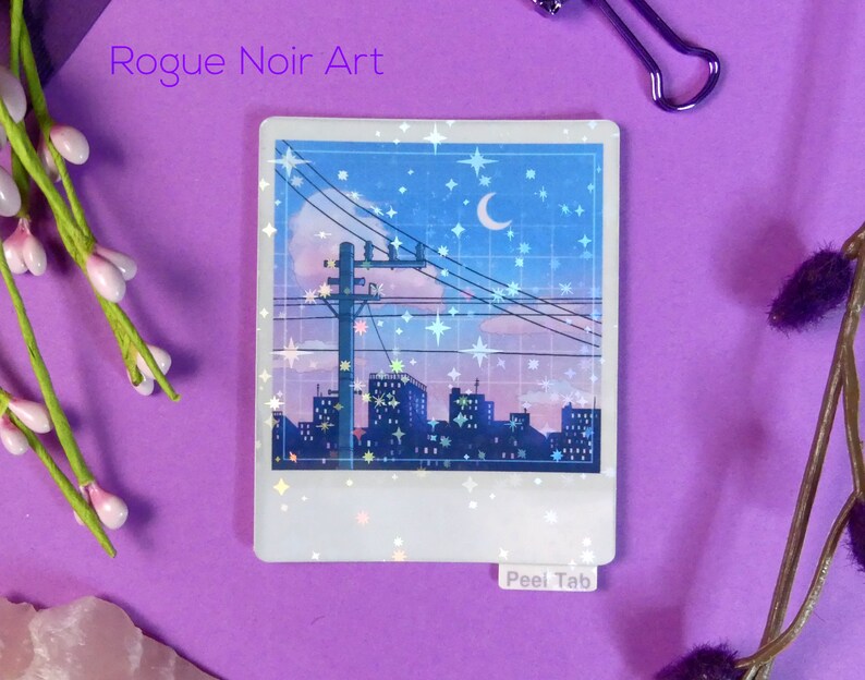 Puede incluir: Una pegatina hologr&aacute;fica que representa una ciudad nocturna con una luna creciente y estrellas. La pegatina tiene un borde blanco y una pesta&ntilde;a de despegue que dice "Peel Tab".