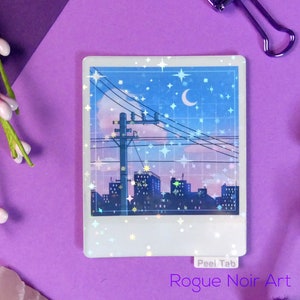 Puede incluir: Una pegatina hologr&aacute;fica con una ciudad nocturna con una luna creciente y estrellas. La pegatina tiene un borde blanco y el texto "Peel Tab" y "Rogue Noir Art" est&aacute;n impresos en la parte inferior.
