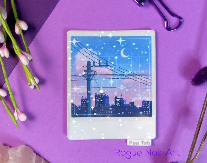 Puede incluir: Una pegatina hologr&aacute;fica con una ciudad nocturna con una luna creciente y estrellas. La pegatina tiene un borde blanco y el texto "Peel Tab" y "Rogue Noir Art" est&aacute;n impresos en la parte inferior.