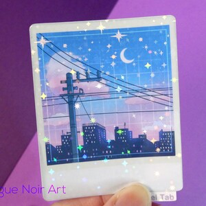 Puede incluir: Una pegatina hologr&aacute;fica con una ciudad nocturna con una luna creciente y estrellas en el cielo. La ciudad est&aacute; silueteada contra un cielo rosa y azul. La pegatina tiene un acabado brillante.