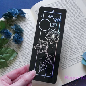 May include: A black bookmark with a holographic finish features a white line drawing of a moon, stars, and a vine with flowers. The bookmark is approximately 10 cm tall and 5 cm wide.