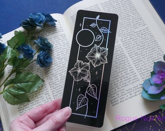 May include: A black bookmark with a holographic finish features a white line drawing of a moon, stars, and a vine with flowers. The bookmark is approximately 10 cm tall and 5 cm wide.