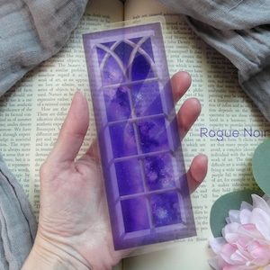 May include: A purple and white stained glass bookmark with a galaxy design. The bookmark is shaped like a tall, narrow rectangle with a pointed top.
