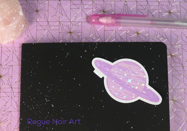 K&ouml;nnte beinhalten: Ein holografischer Aufkleber eines lila Planeten mit einem Ring darum auf einem schwarzen Hintergrund mit wei&szlig;em Glitzer. Der Aufkleber sagt "Rogue Noir Art" in wei&szlig;er Schrift.