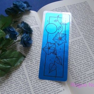 May include: A blue and black bookmark with a floral design and a moon. The bookmark is made of a shiny, metallic material.