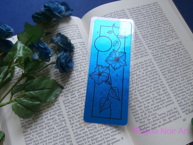 May include: A blue and black bookmark with a floral design and a moon. The bookmark is made of a shiny, metallic material.
