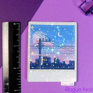 Puede incluir: Una pegatina hologr&aacute;fica que representa una ciudad nocturna con una luna creciente y estrellas en el cielo. La pegatina tiene un borde blanco y una pesta&ntilde;a de despegue que dice "Peel Tab". El texto "Rogue Noir Art" est&aacute; impreso debajo de la pegatina.