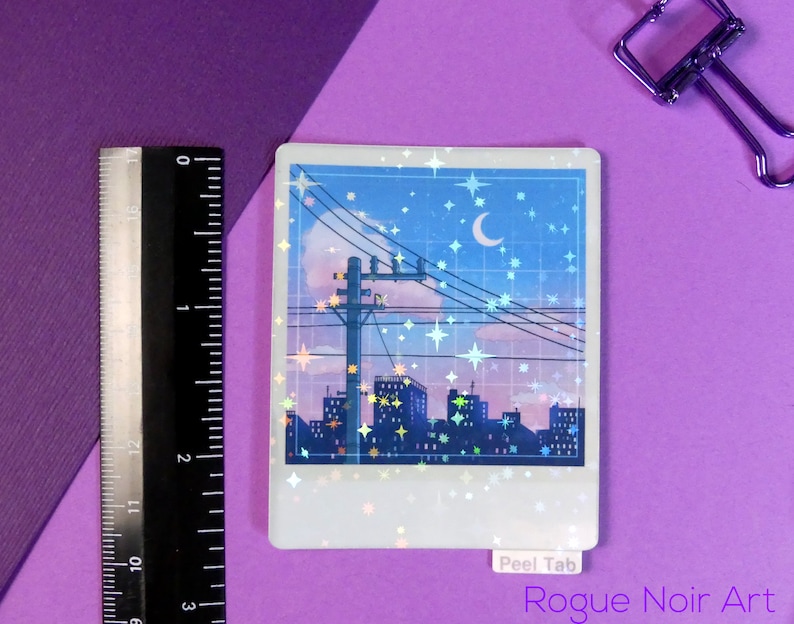 Puede incluir: Una pegatina hologr&aacute;fica que representa una ciudad nocturna con una luna creciente y estrellas en el cielo. La pegatina tiene un borde blanco y una pesta&ntilde;a de despegue que dice "Peel Tab". El texto "Rogue Noir Art" est&aacute; impreso debajo de la pegatina.