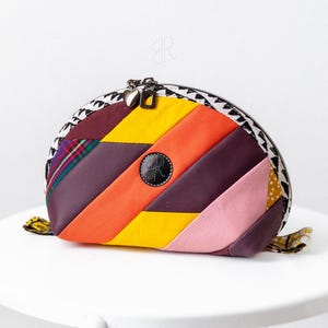 May include: A vibrant, patchwork zipped pouch. The pouch is made from various fabrics in shades of purple, orange, yellow, and pink, arranged in diagonal stripes. A black button with a design is centered on the front. The zipper pulls are heart-shaped.