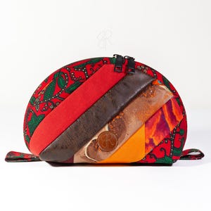 Pode incluir: Uma clutch em forma de meia-lua com uma base de tecido vermelho estampado. O design apresenta listras diagonais vermelhas, marrons, laranja e tecido estampado. Um pequeno círculo de couro com um logotipo está na frente. Possui um zíper preto.