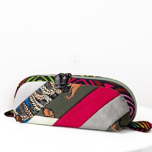 Può includere: Un astuccio colorato con un design patchwork. L'astuccio presenta una varietà di tessuti nei toni del verde, rosa, grigio e tessuti fantasia. Ha una cerniera nera e un piccolo anello su un'estremità.