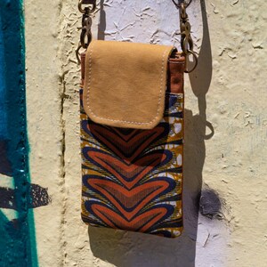 Könnte beinhalten: Eine kleine, rechteckige Handytasche mit Klappenverschluss. Die Tasche hat ein farbenfrohes geometrisches Muster in Orange, Blau und Gelb. Die Klappe und der obere Teil sind einfarbig beige. Sie hängt an einer bronzefarbenen Kette.