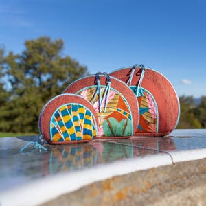 Peut inclure: Trois pochettes zippées rondes en forme de demi-lune avec des motifs floraux et géométriques colorés. Les pochettes sont en tissu tissé avec une bordure en cuir marron. Les pochettes sont imbriquées les unes dans les autres, la plus grande en bas et la plus petite en haut.