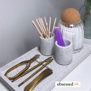 May include: A white tray with a speckled finish holds various nail care tools.  The tools include a pair of gold nail clippers, a gold cuticle pusher, a gold nail file, and a pair of gold tweezers.  There are also two small white speckled containers, one holding wooden cuticle sticks and the other holding purple cotton swabs.  A clear glass jar with a cork lid holds cotton balls.  The tray has the text "obsessed" and "@gold_dust_designs" on it.
