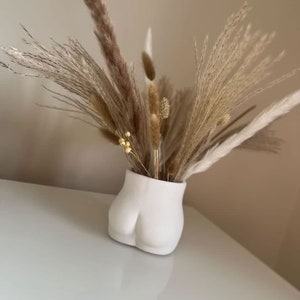 May include: A white ceramic vase shaped like a human bottom, filled with dried pampas grass.
