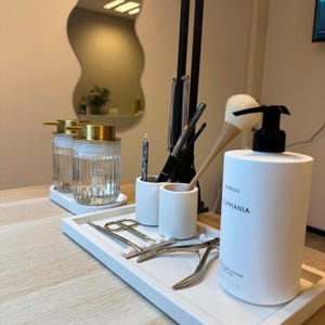 May include: A white tray displays manicure tools, including scissors and clippers, next to a white bottle of hand soap with the text "BYREDO LIPMANIA." Two glass jars with gold lids and a mirror are in the background.