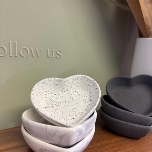 May include: A collection of heart-shaped dishes in shades of white, grey, and black. The top dish is white with black flecks, while others have a marbled effect or are solid colours. These small dishes are ideal for holding jewellery or other small items.