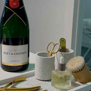 May include: A white tray with a speckled pattern, featuring a bottle of Moët & Chandon champagne, a small bottle of cuticle essence, a pair of gold nail clippers, two gold cuticle pushers, a pair of gold scissors, a small white speckled cup, and a wooden brush with white bristles.